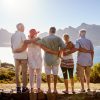 4 friends all over 60 are standing and having a group hug. Their backs are turned to the camera as they look out at a lake view. Sunny day. Friend holiday. Traveling in retirement.